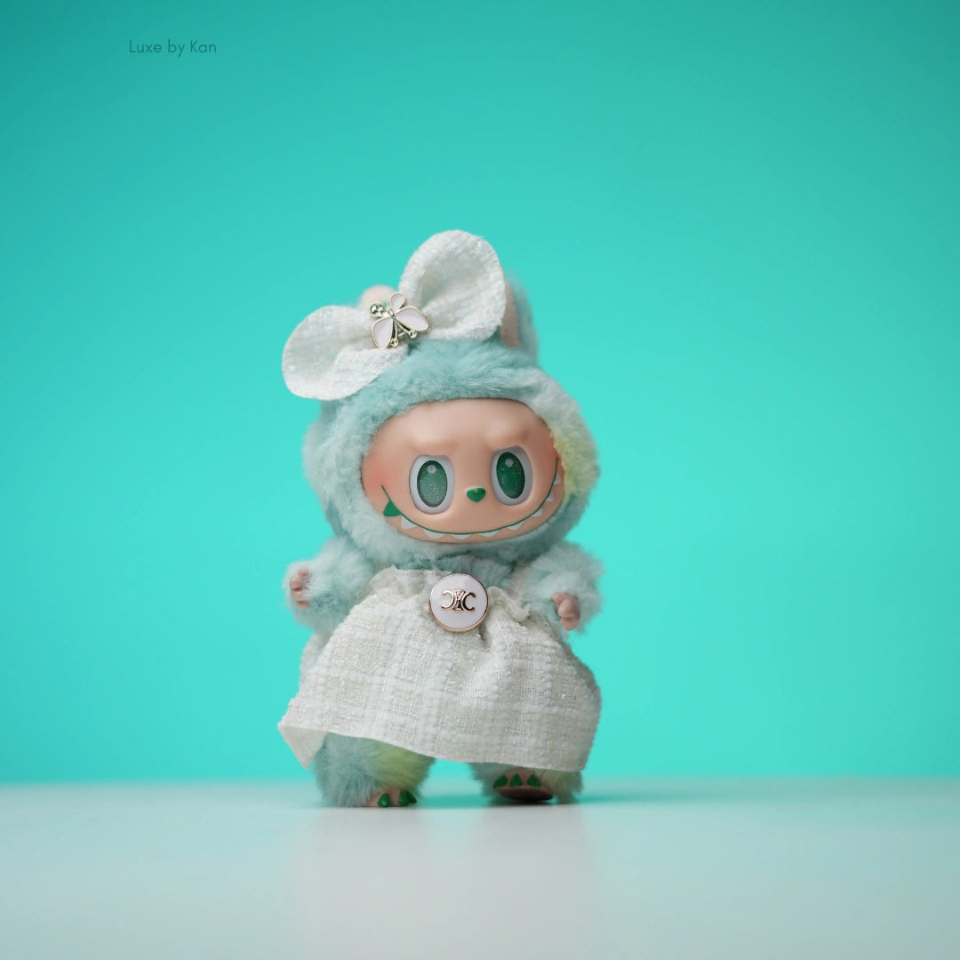 Pop Mart Labubu doll wearing a tweed dress, adorned with a bow. The doll has green eyes, pointy ears and teal fluffy fur.