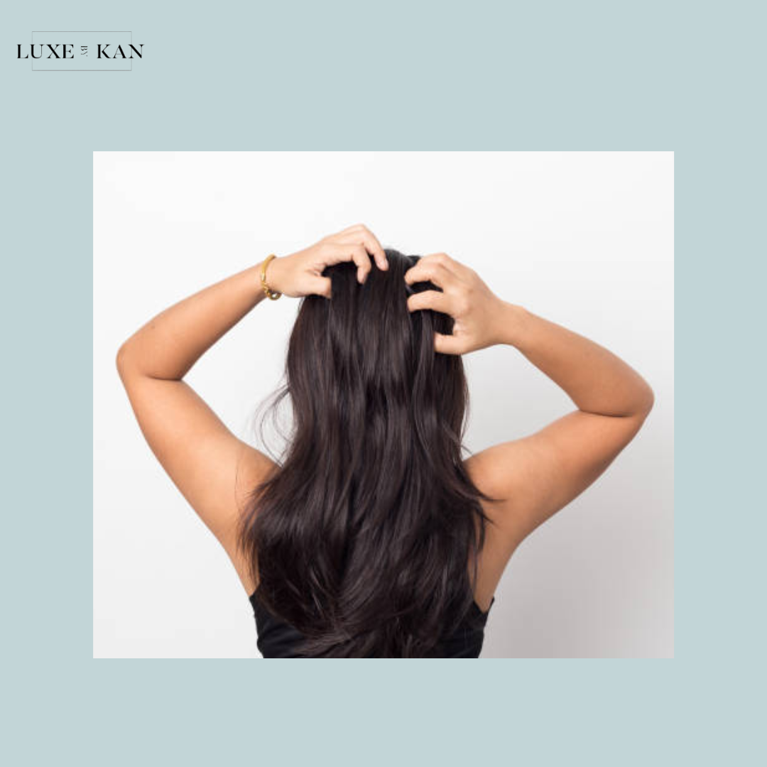 A woman with long, dark hair is shown from behind, gently massaging her scalp with both hands against a light backdrop.