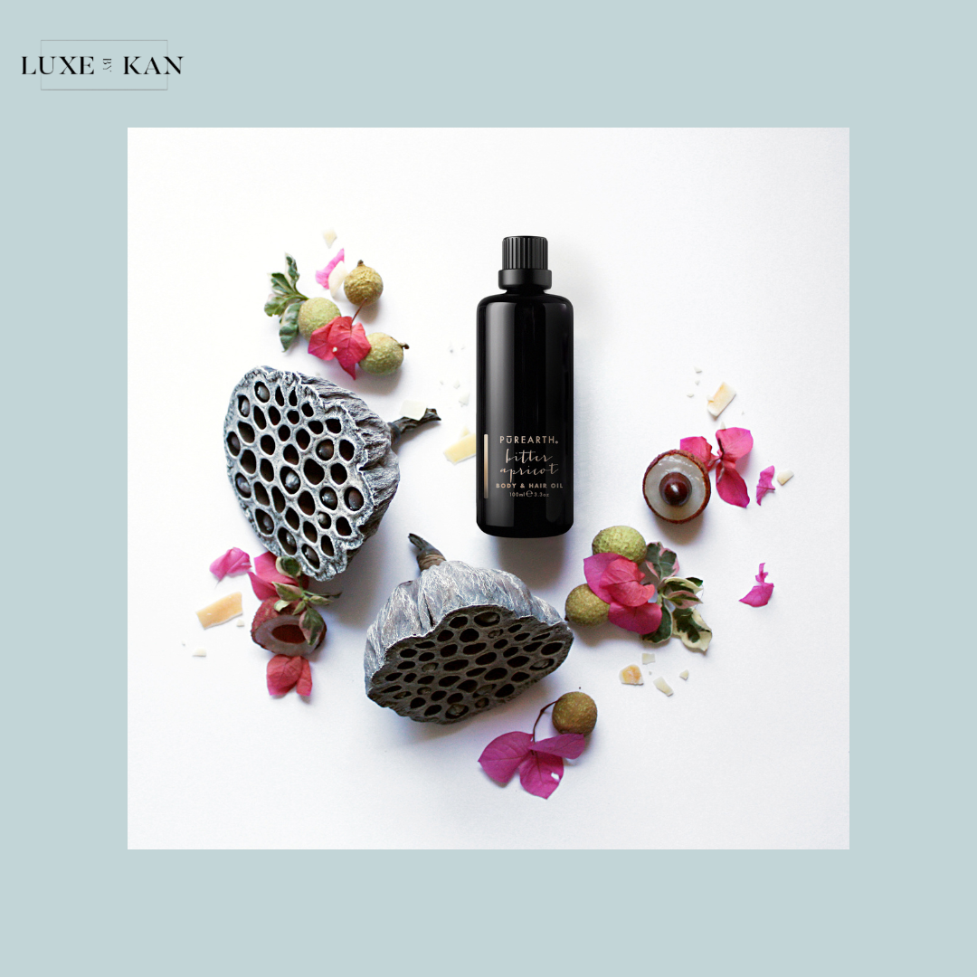 A black bottle labeled "Pure Earth Body & Hair Oil" surrounded by dried lotus pods and colorful petals on a white surface.