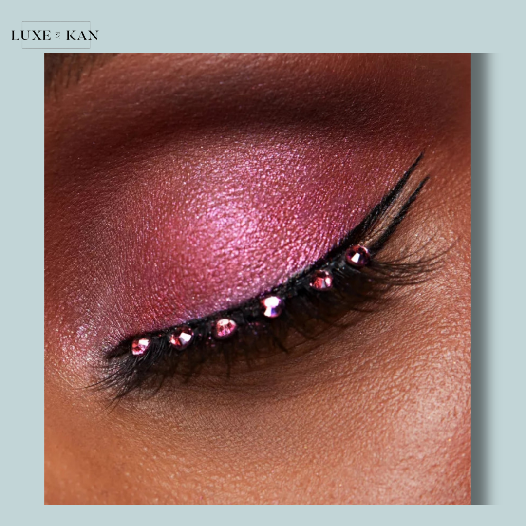 Close-up of an eye with shimmering pink eyeshadow, bold black eyeliner, and decorative pink rhinestones along the lower lash line.