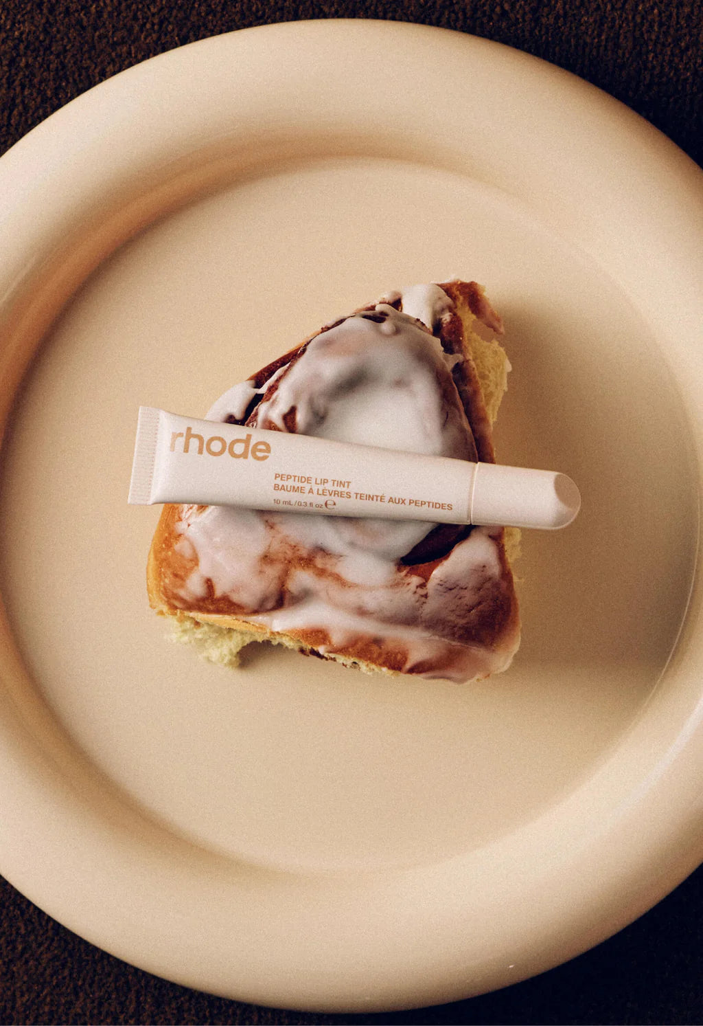Rhode Peptide Lip Treatment Cinnamon Roll on a beige plate with a cinnamon roll slice topped with white frosting.