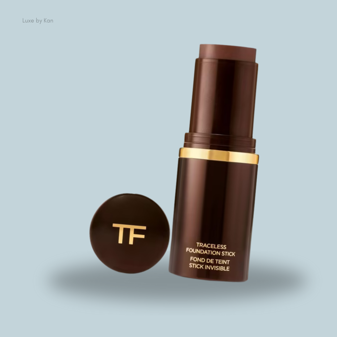 Brown foundation stick with a gold band and "TF" logo, accompanied by a round compact, set against a light blue background.