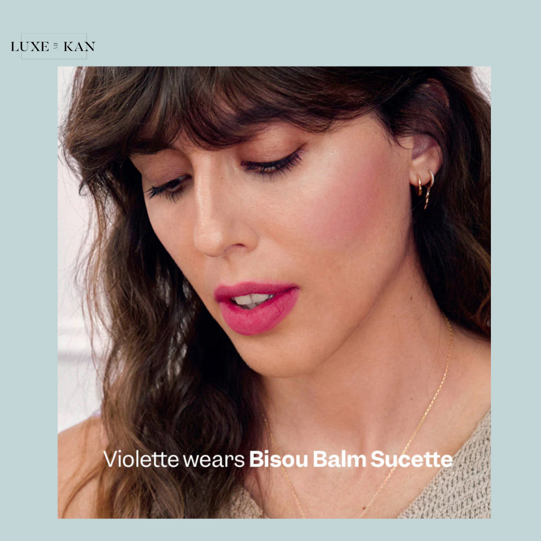 A close-up of a woman with wavy hair wearing subtle jewelry, showcasing the Bisou Balm Suquette product.