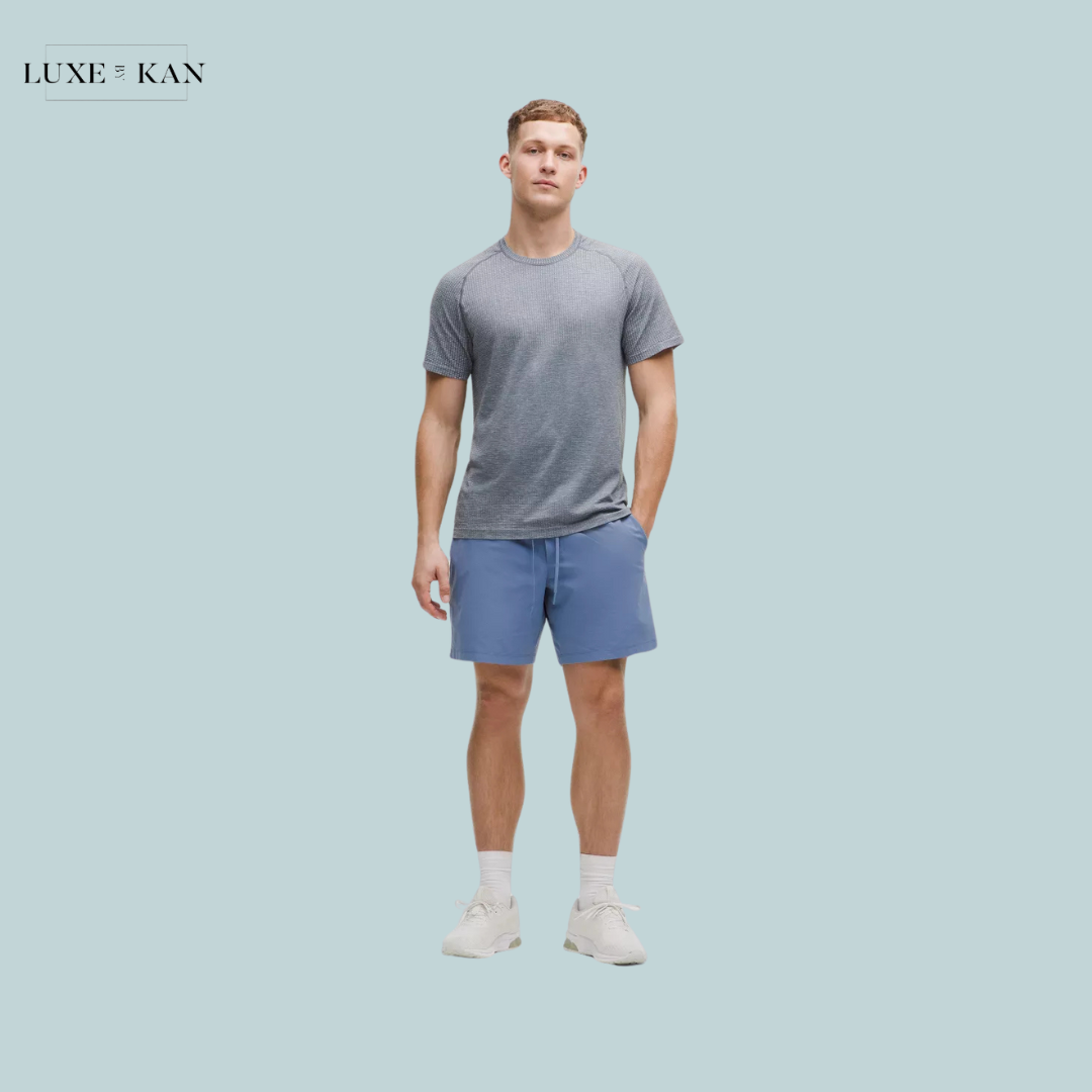 A model in a gray athletic t-shirt and blue shorts stands against a light blue background, showcasing stylish activewear attire.