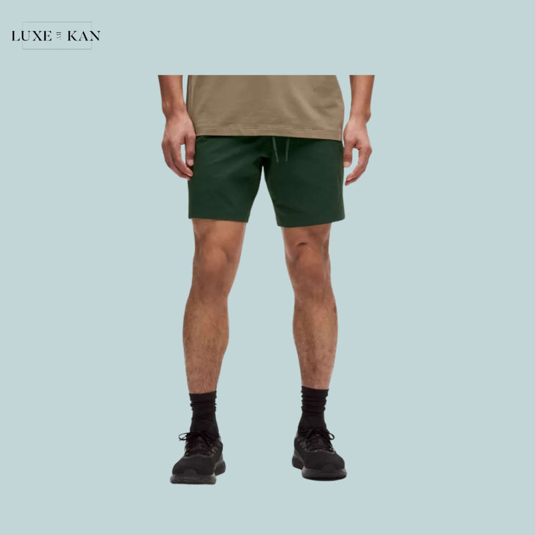 A man stands wearing green shorts and a beige shirt, paired with black socks and shoes.