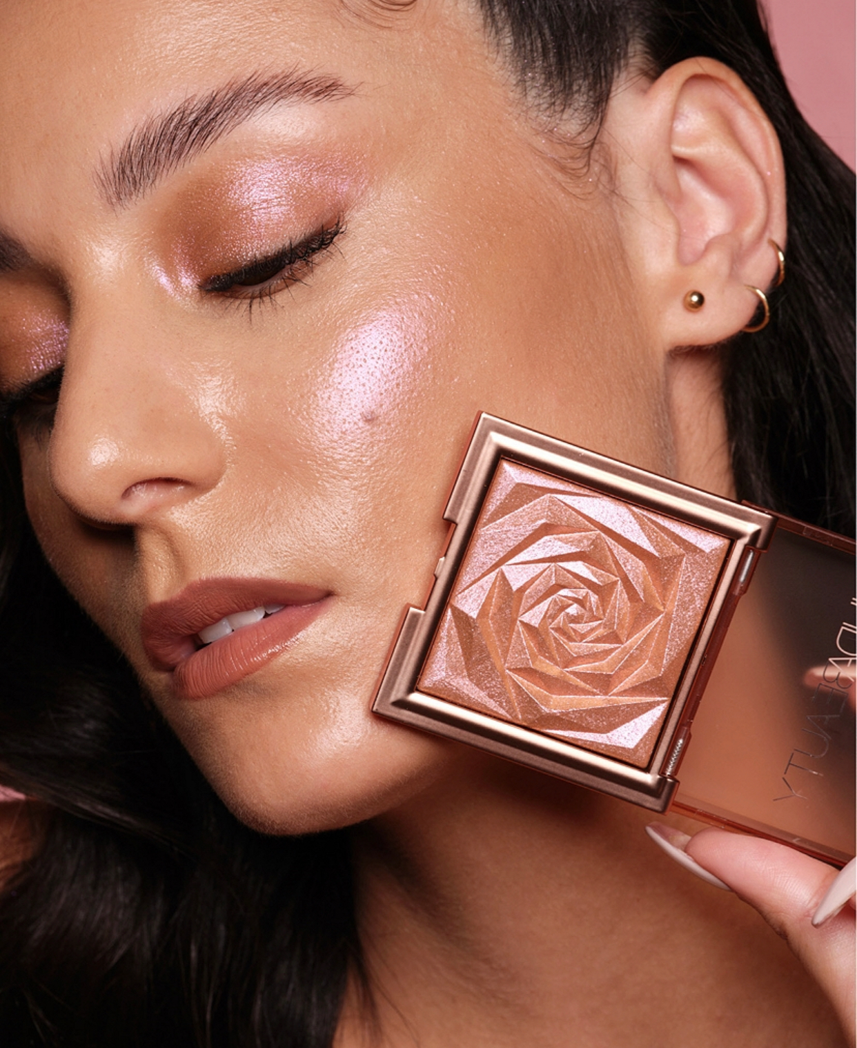 A close-up of a woman's ear with multiple earrings, holding a bronzer compact featuring geometric patterns and a shiny finish.