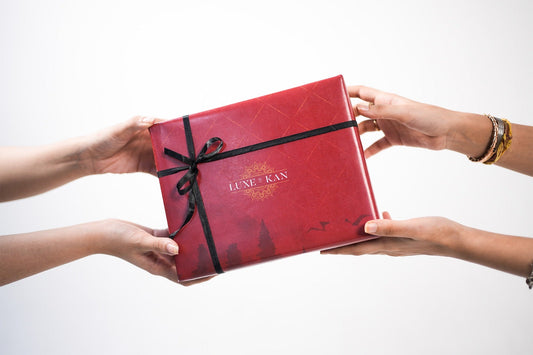 Red "Luxe By Kan" branded gift box with a black ribbon being exchanged between two sets of hands against a white backdrop.
