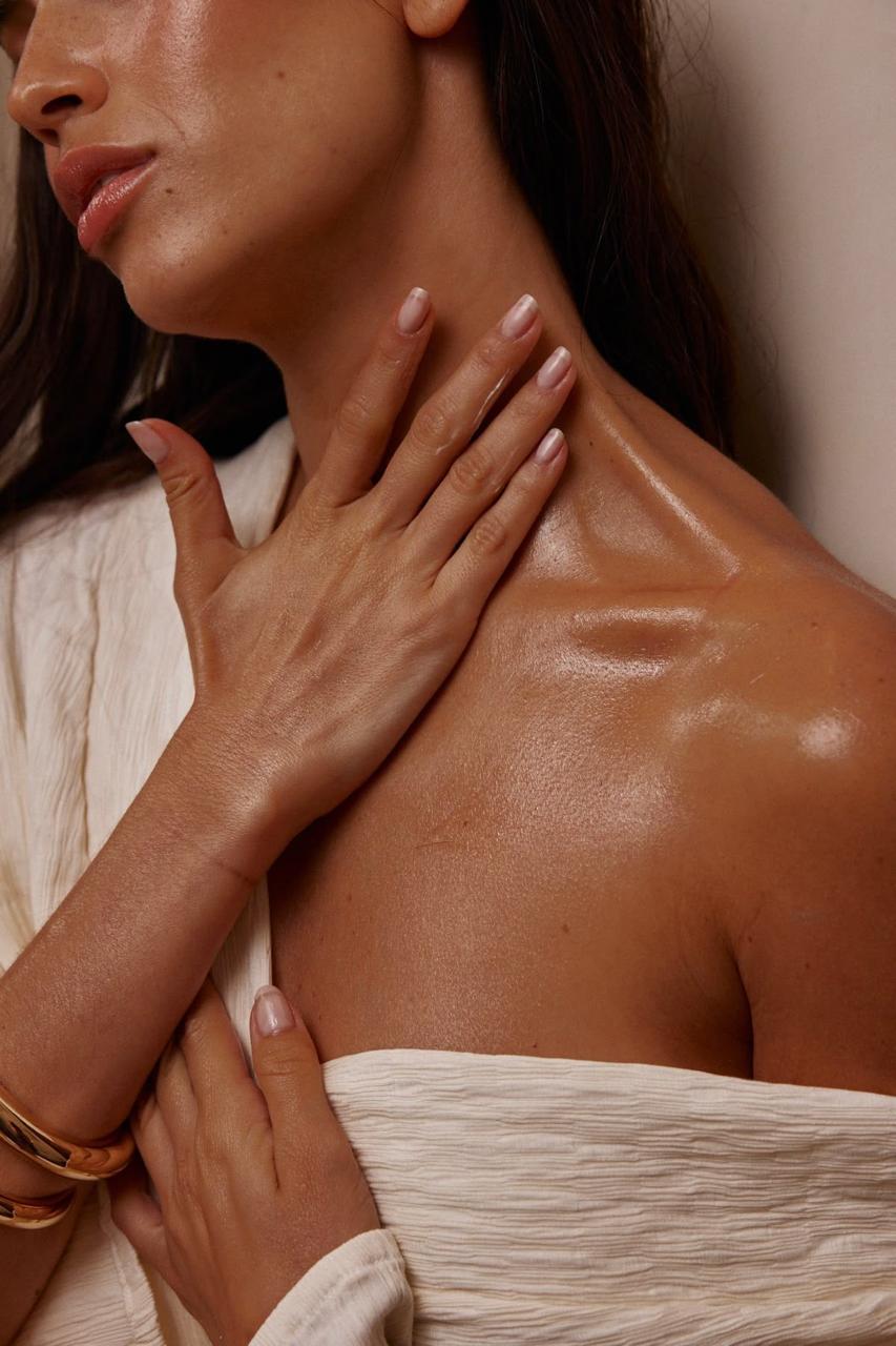 A woman with glowing, tan skin wears a white robe and touches her neck, showcasing her shimmering chest and collarbone.