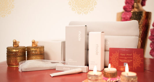 Self-care gift hamper including containers, candles, beauty products from Rhode, and a Diwali greeting card on a red surface.