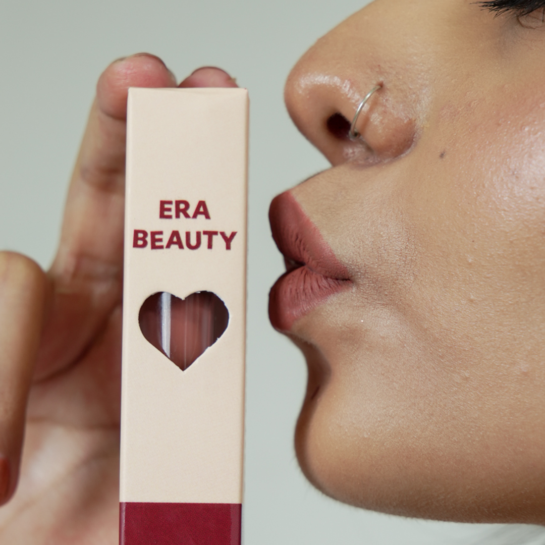 Close-up of a person's face wearing matte lipstick along with Era Beauty product packaging showing through a heart cutout.