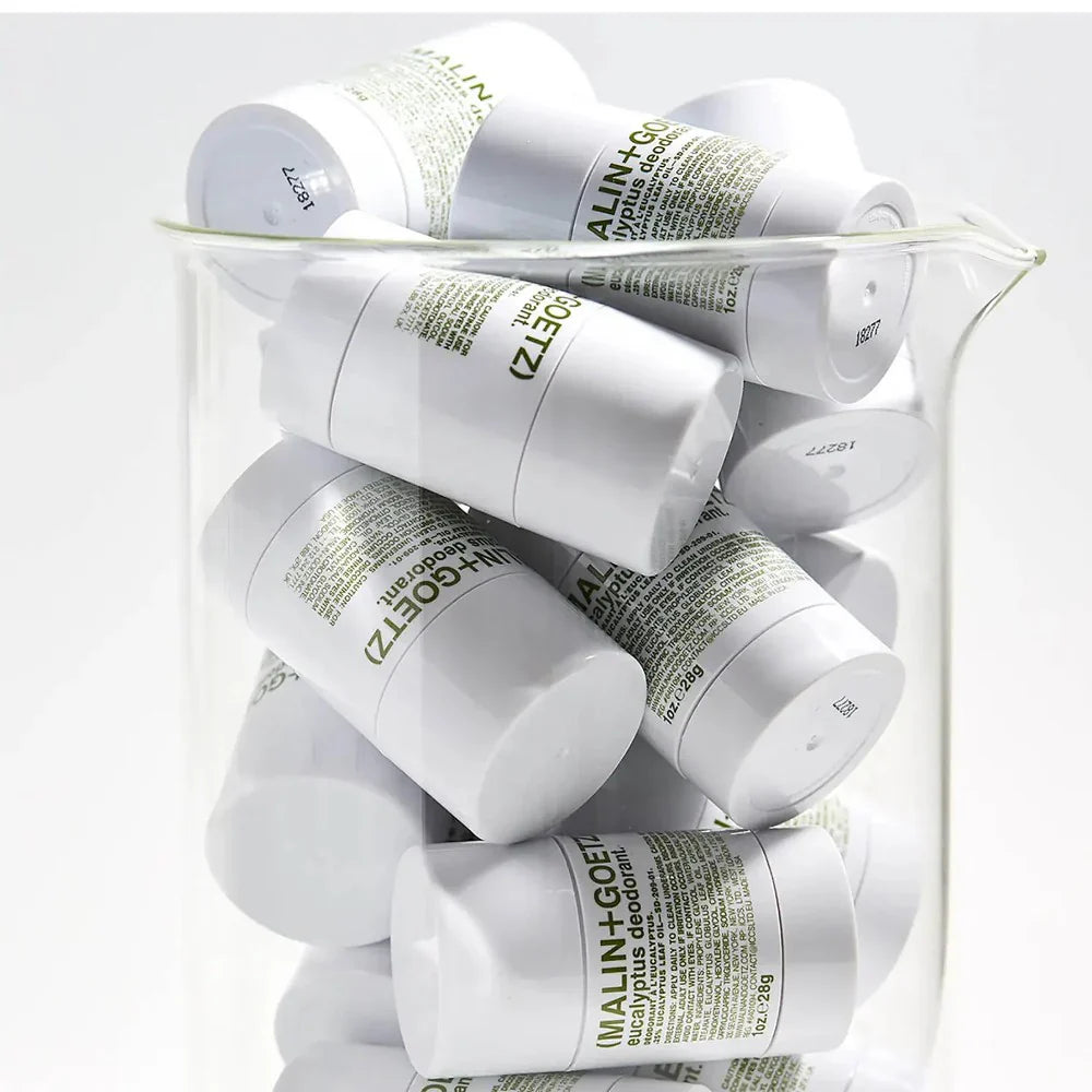 Several white sticks of Malin+Goetz Eucalyptus Deodorant are piled inside a clear glass lab beaker against a white wall.