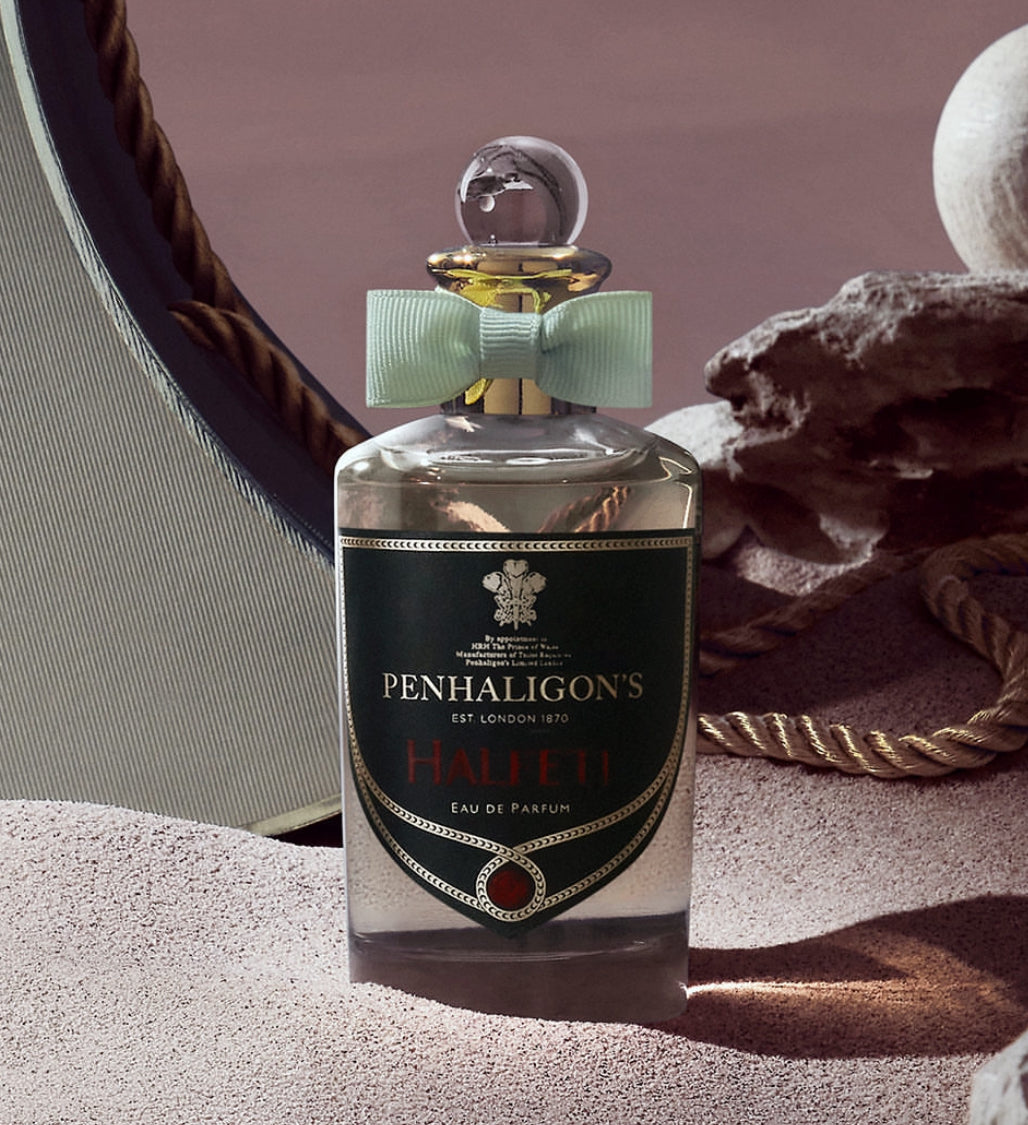 Penhaligon's 'Haleti' Eau de Parfum, clear glass bottle with green bow on sand, alongside rocks and other props.