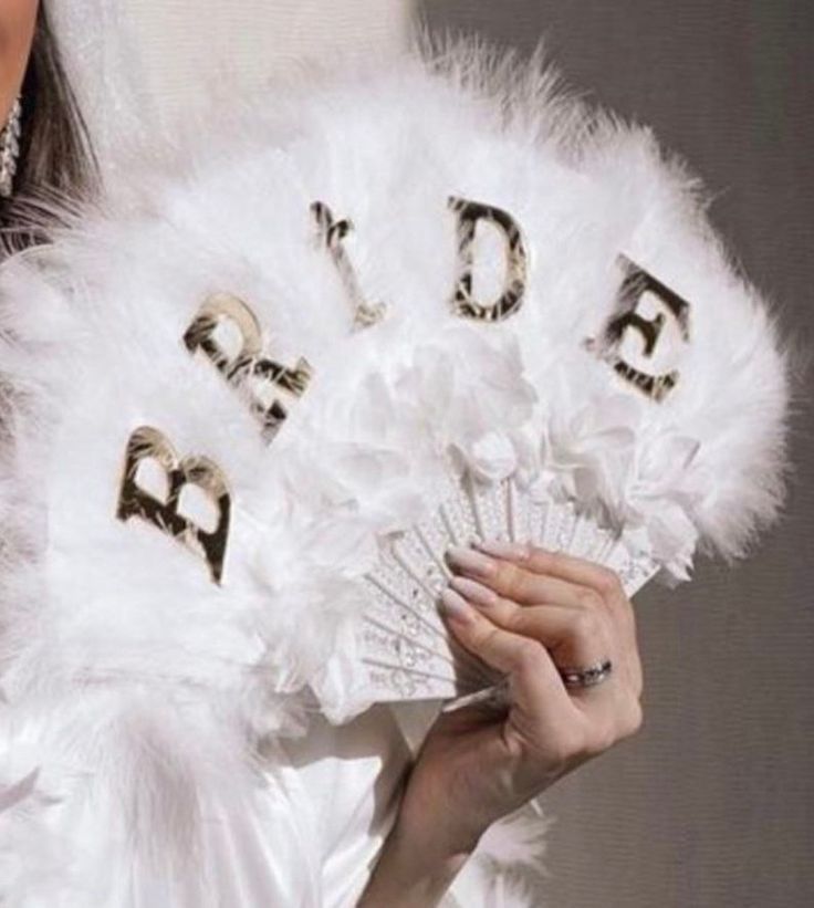 White feather fan with golden letters that spell "BRIDE," held by a hand with light pink nails and a ring on the ring finger.