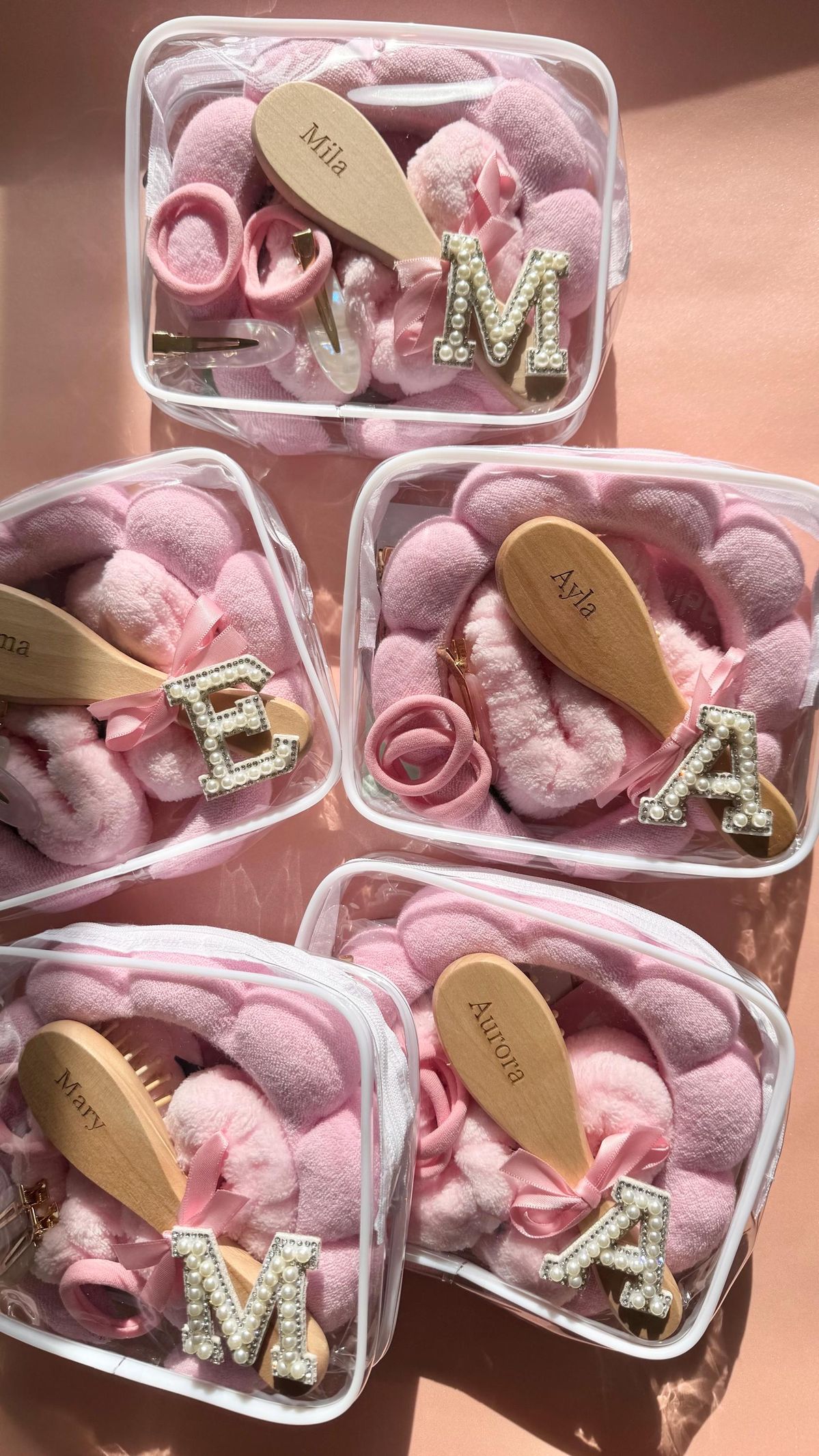 Five clear toiletry bags filled with pink accessories, each labelled with the bridesmaids' names and their first initial.