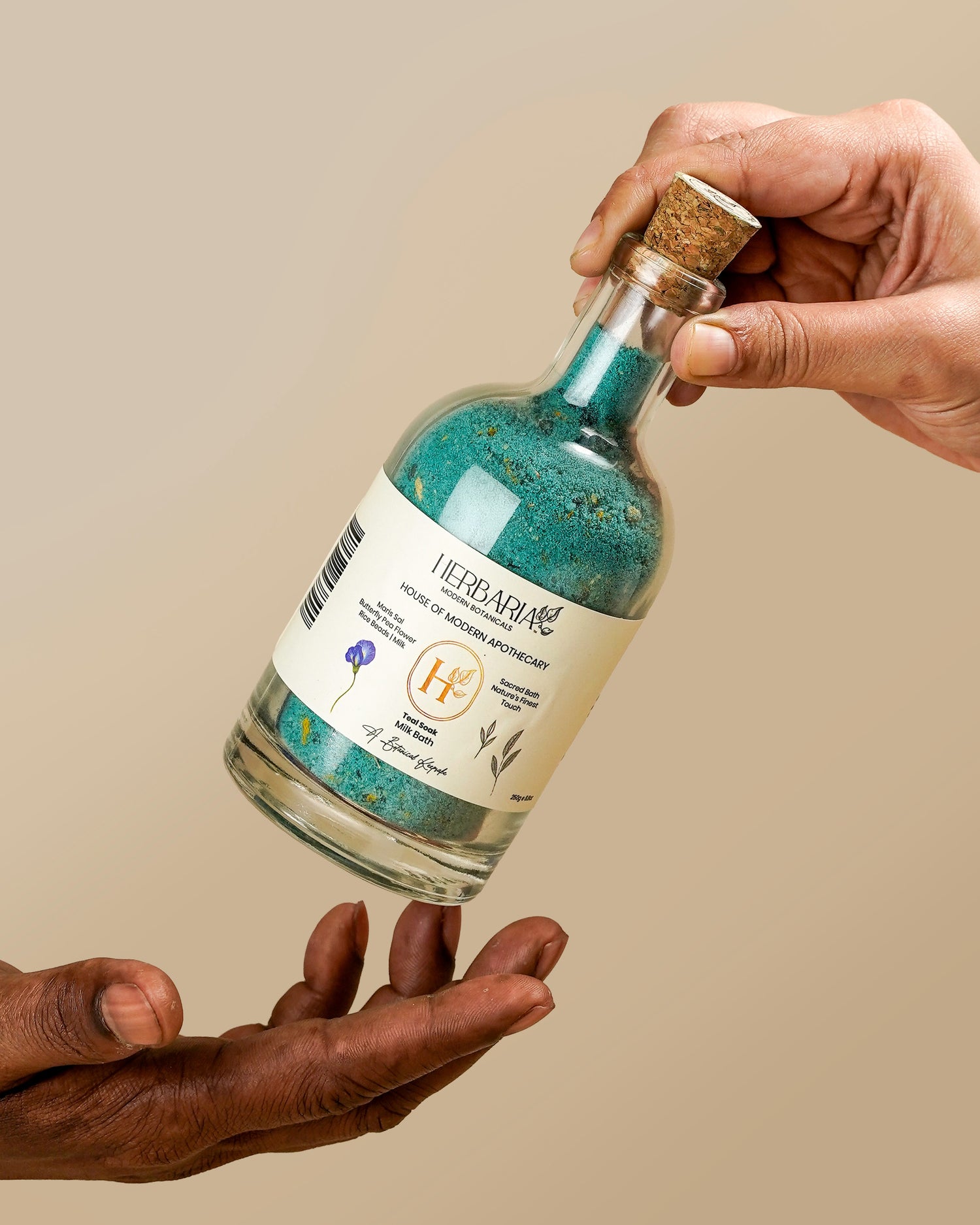 Hands holding a glass bottle of Herbaria Teal Soak milk bath salts with a cork stopper against a tan background.