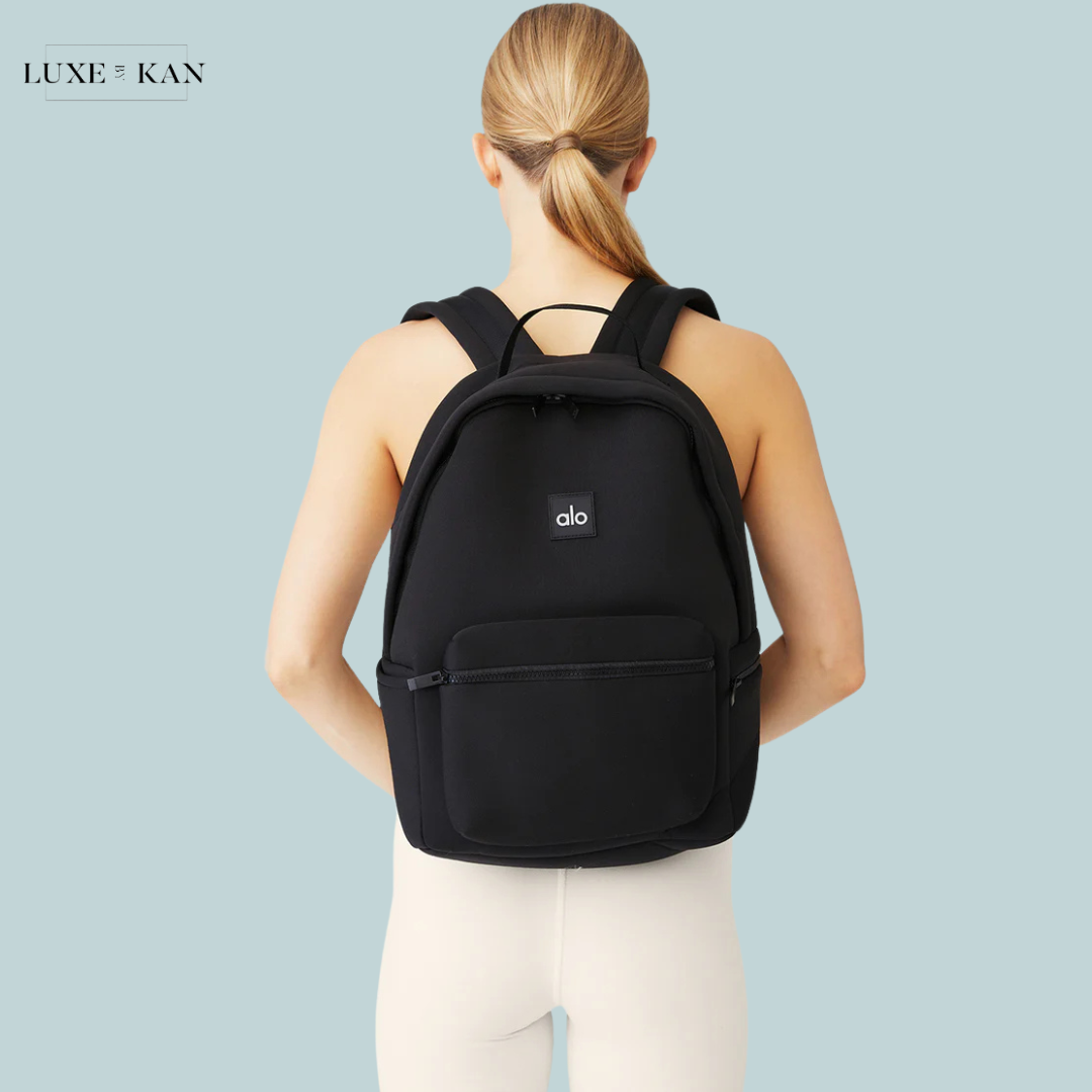 A woman in a black Alo backpack stands facing away against a light blue background, showcasing the backpack's sleek design.