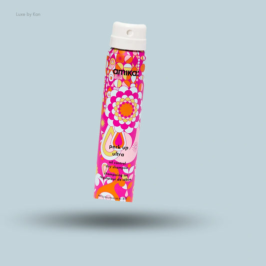 Amika Perk Up Ultra oil control dry shampoo in a vibrant pink patterned spray bottle against a light blue background.