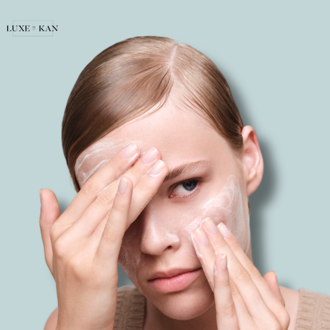 A woman with sleek, light brown hair applies skincare, set against a soft blue background, wearing a knitted top.