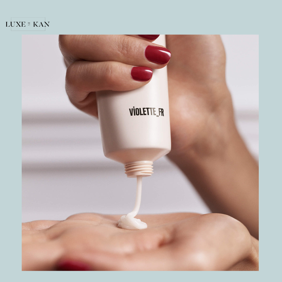 A hand with red nails squeezes a c'est la moisturising cream tube, dispensing a dollop of white lotion onto an open palm.