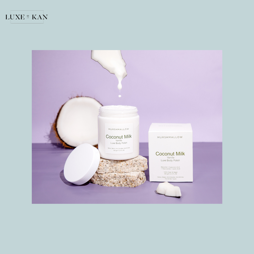 A jar of Coconut Milk Luxe Body Polish, surrounded by coconut halves and a box, with cream drizzling from the lid.