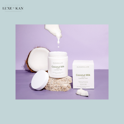 A jar of Coconut Milk Luxe Body Polish, surrounded by coconut halves and a box, with cream drizzling from the lid.