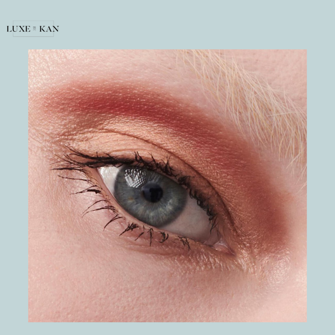 Close-up of a blue eye with warm peach and copper eyeshadow, showcasing defined lashes and subtle blonde eyebrows.