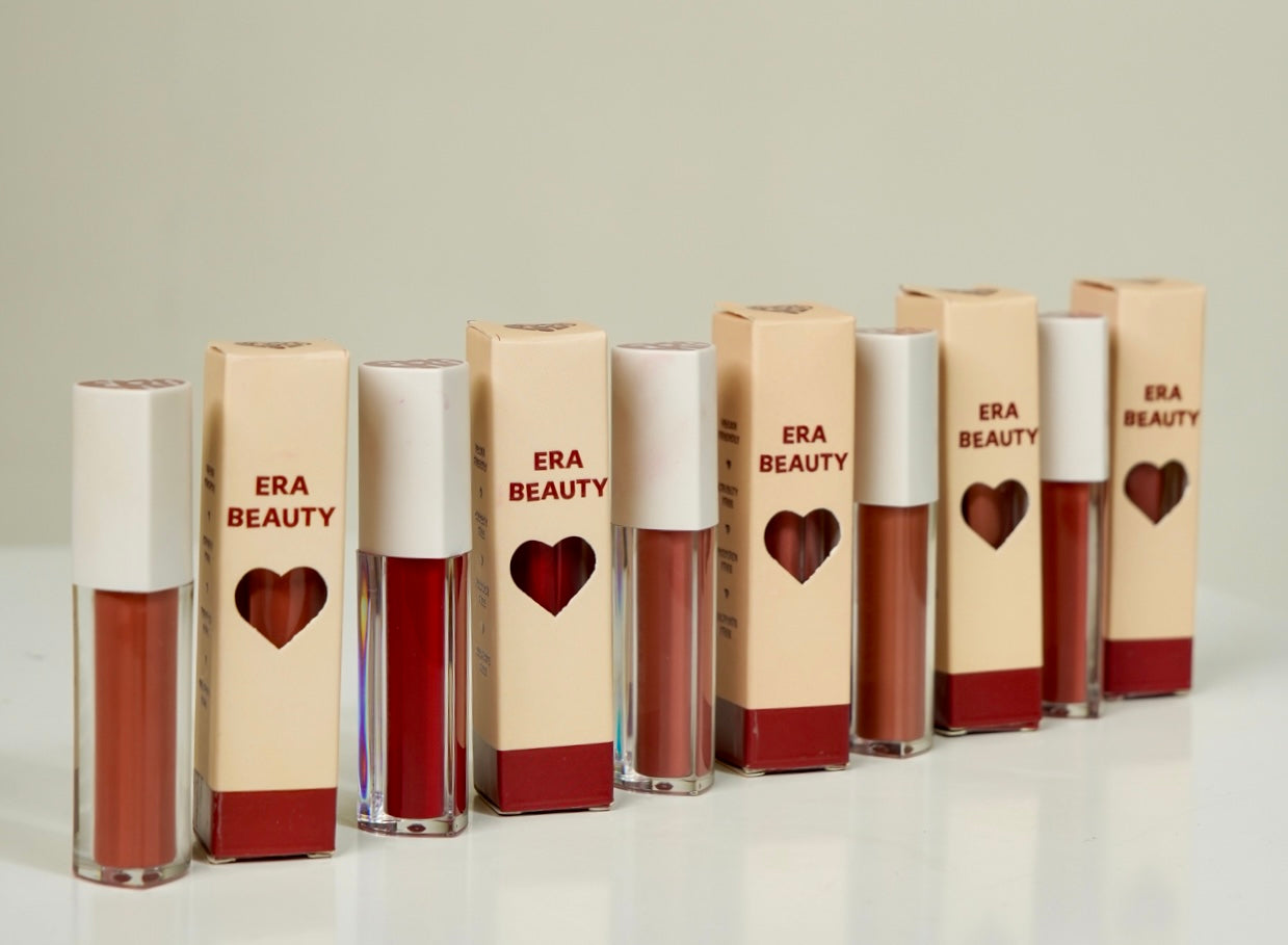Row of Era Beauty liquid lipsticks in various shades, packaged in beige boxes with heart cutouts, displayed on a white surface.
