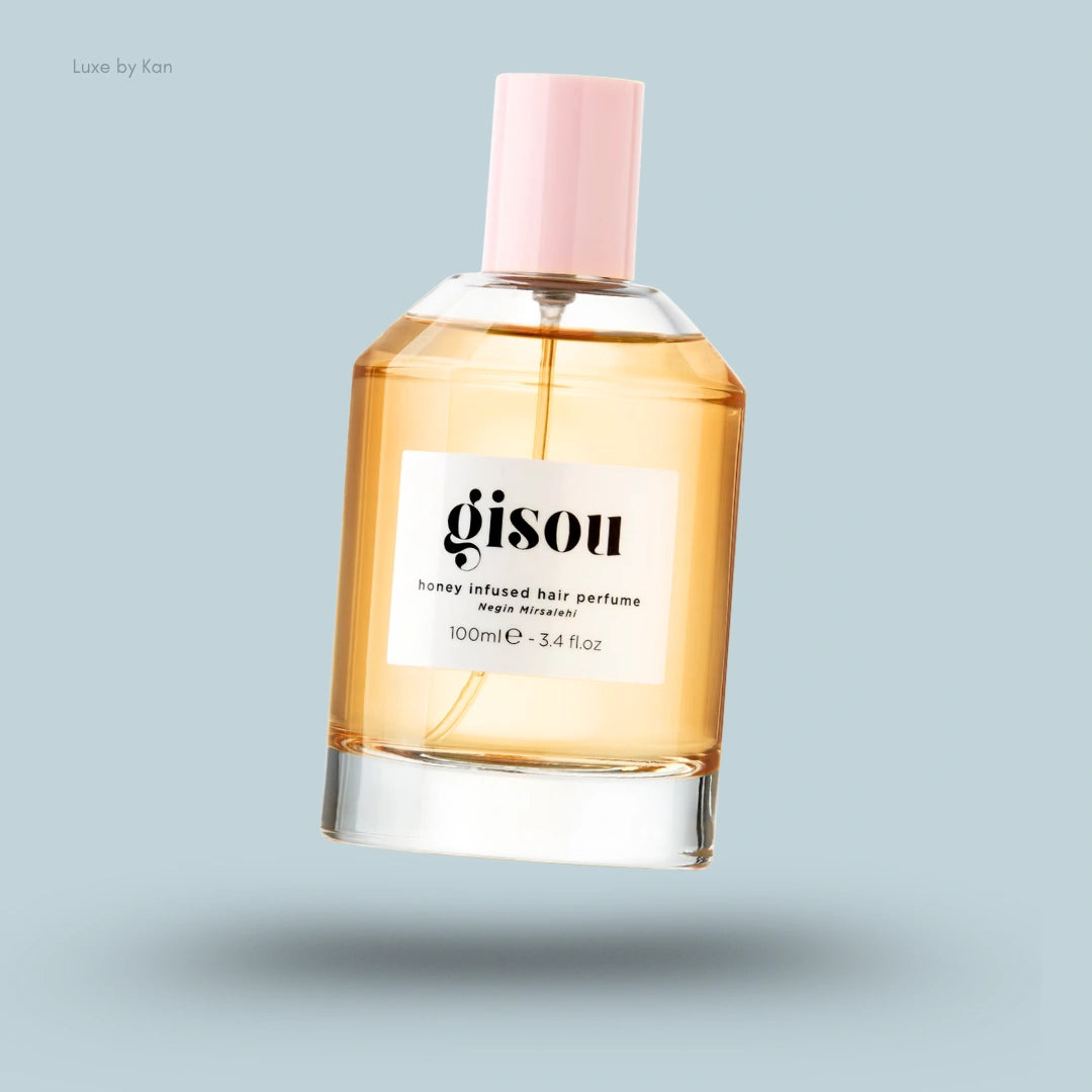 A clear glass bottle of Gisou Honey Infused Hair Perfume with a light amber liquid and a pink cap on a pale blue background.