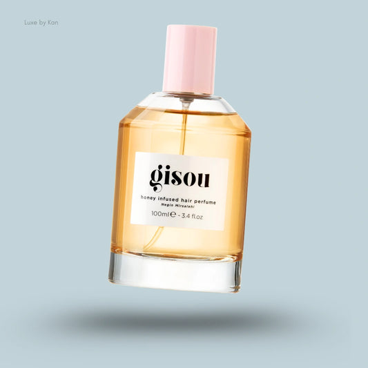 A clear glass bottle of Gisou Honey Infused Hair Perfume with a light amber liquid and a pink cap on a pale blue background.