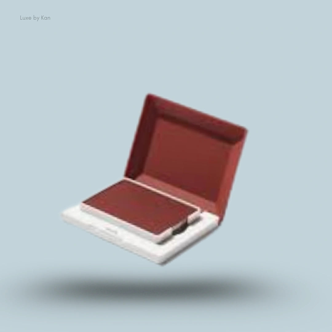 An open compact case with Glossier Cloud Paint Plush Blush in a deep reddish-brown shade on a light blue background.