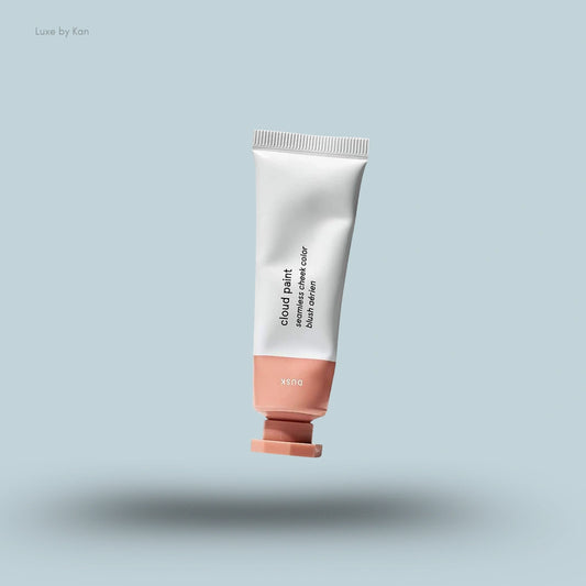 A tube of Glossier Cloud Paint Seamless Cheek Color in the shade "Dusk" floats against a light blue background.