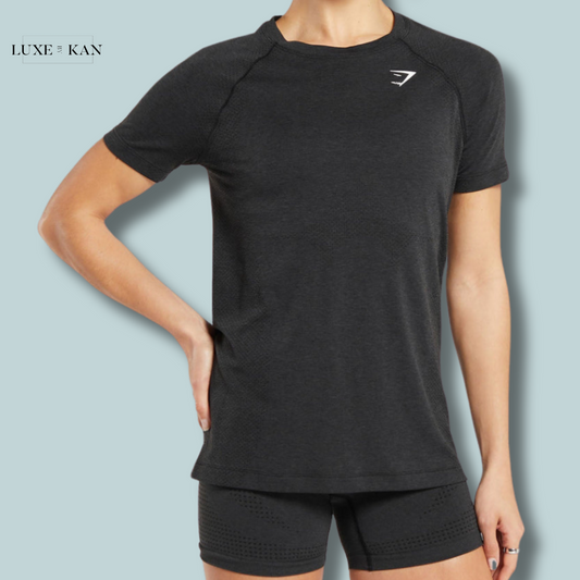 Woman wearing a stylish black gymshark vital seamless light t-shirt and shorts, posing confidently against a soft blue background. The logo is visible on the shirt.