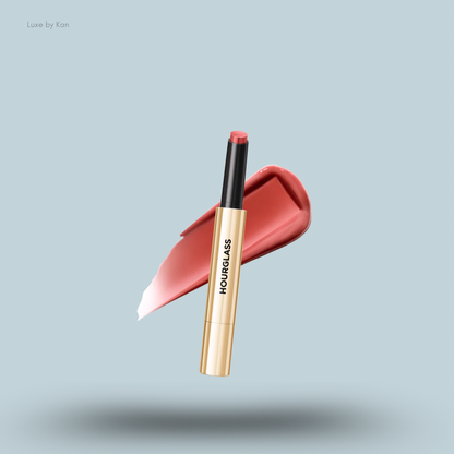 A gold and black Hourglass Phantom Volumizing Glossy Lip Balm tube is placed over a glossy pink lip product swatch.