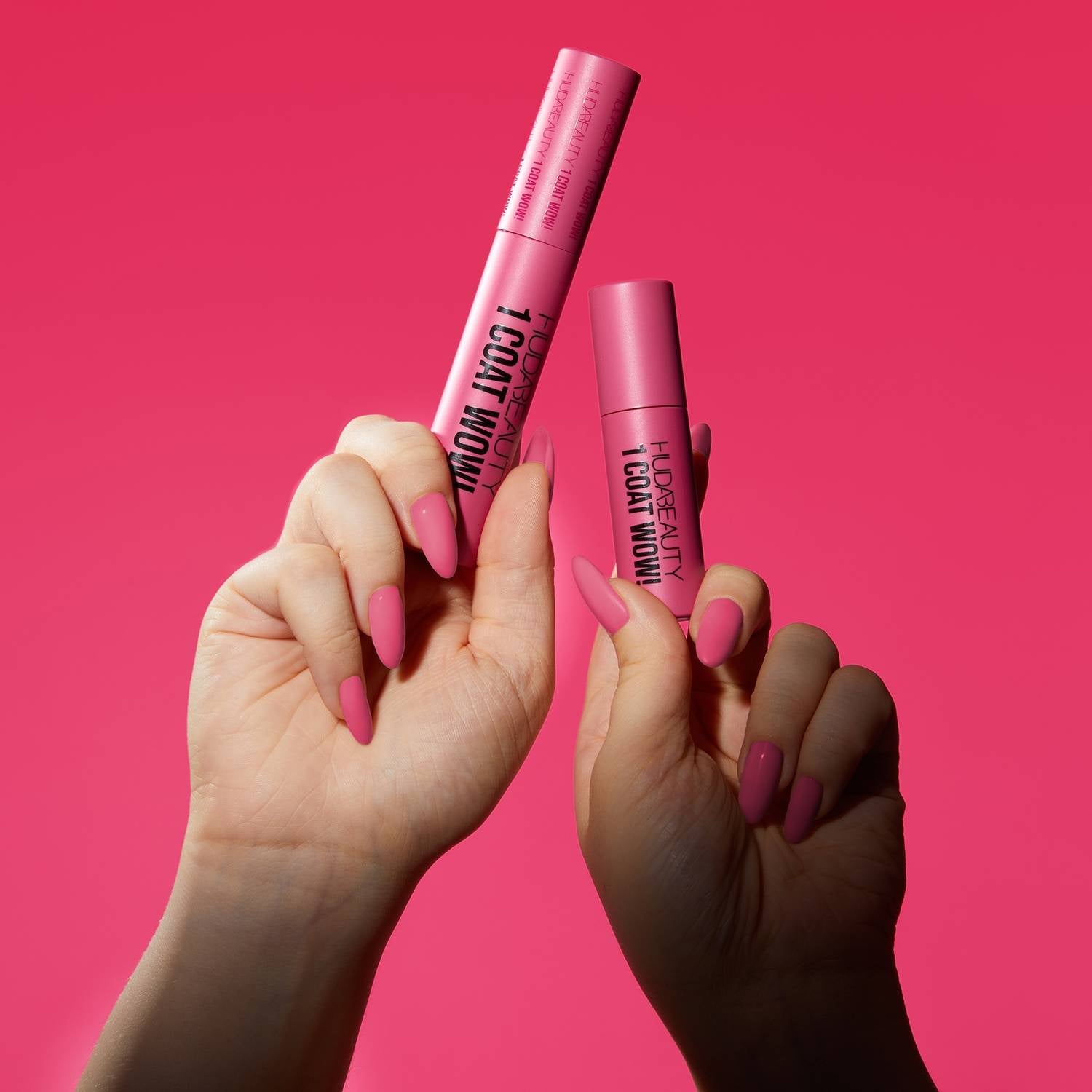 Two hands hold pink huda beauty 1 coat wow! Extra volumising and lifting mascara tubes against a vibrant pink background, showcasing bold beauty in a stylish pose.