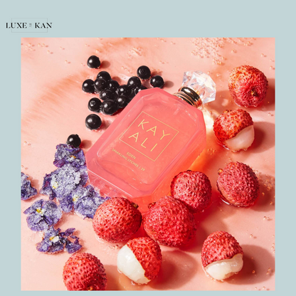 A pink perfume bottle labeled "KAYALI EDEN" surrounded by lychee fruit, blueberries, and violet petals on a pale pink surface.