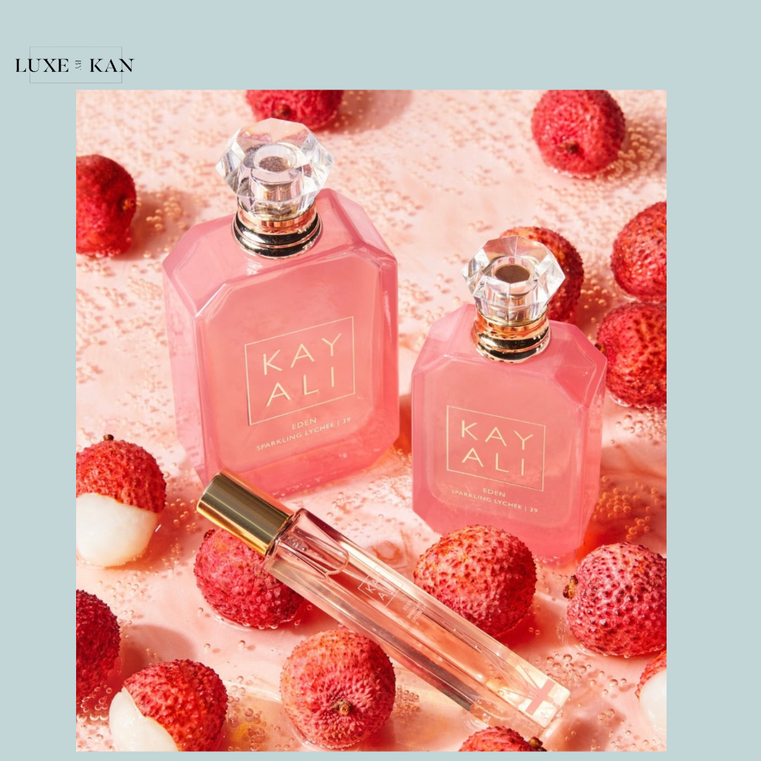 Three pink perfume bottles labeled "KAYALI" sit atop a pink surface dotted with lychee fruit, evoking a fresh, fruity aesthetic.