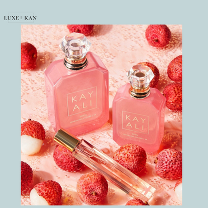 Three pink perfume bottles labeled "KAYALI" sit atop a pink surface dotted with lychee fruit, evoking a fresh, fruity aesthetic.
