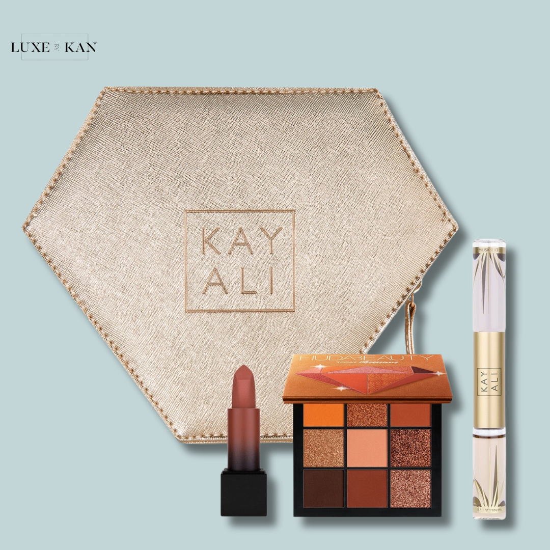 A hexagonal, gold makeup pouch containing a mauve lipstick, an eyeshadow palette with warm tones, and a clear lip gloss, all branded Kayali.