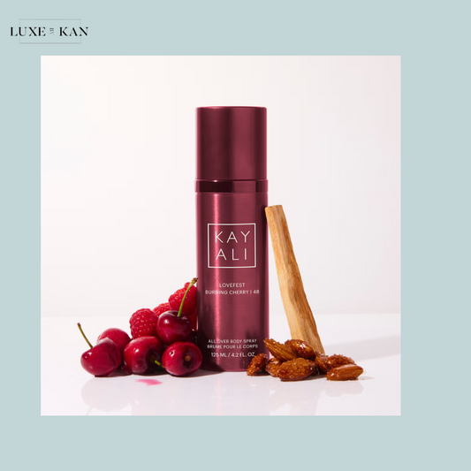 A burgundy KAYALI Lovefest Burning Cherry body spray bottle surrounded by cherries, raspberries, almonds, and a wooden stick.