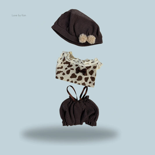 Labubu cocoa cuddle 3-piece set includes a cocoa-colored knit hat with pom poms, a cow-print top with lace trim and a brown bow, and cocoa-colored bloomers.