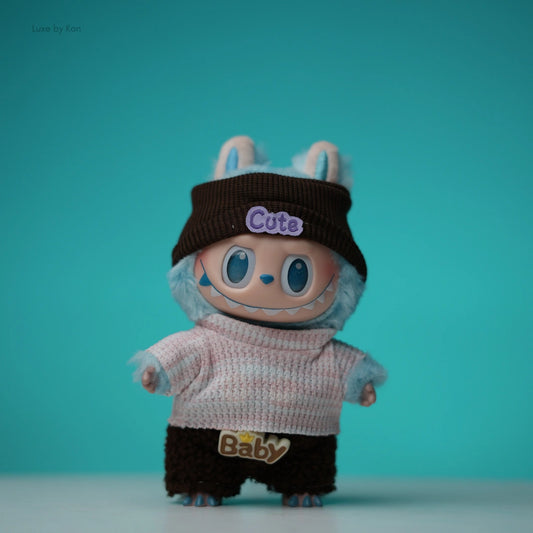 Labubu doll dressed in a brown beanie with “Cute” embroidered on it, a pink and white sweater, and brown shorts featuring the word “Baby.”