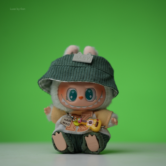 The Labubu doll is dressed in a little explorer three-piece set, complete with a green corduroy bucket hat, a fluffy green-and-white outfit, and a tiny yellow ukulele.