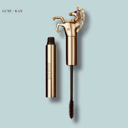 Gold Lash lasso lash wrap mascara tube with a horse-shaped cap adorned with gems, resting beside a black mascara wand on a light blue background.