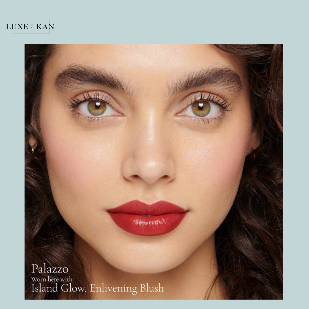 A close-up of a model with curly hair showcasing makeup with labels for "Island Glow" and "Enlivening Blush" below.