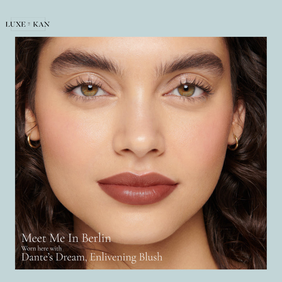 Close-up of a model's shoulder-length wavy hair and gold hoop earrings, featuring the product label "Dante’s Dream, Enlivening Blush."