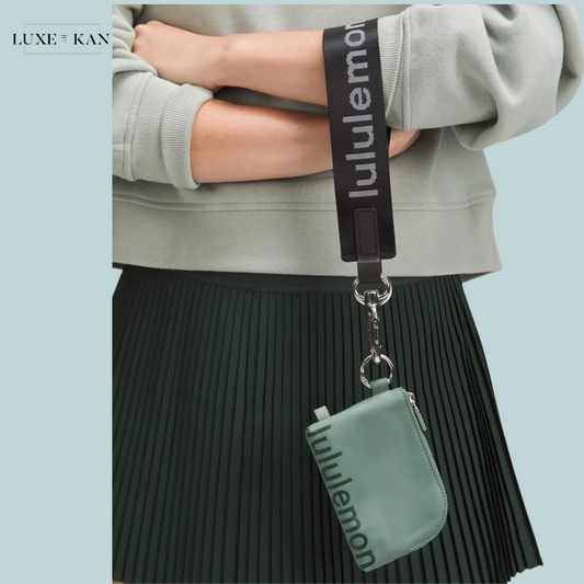 A close-up of a person's arm wearing a light green sweatshirt, holding a lanyard with a mint green clippable card pouch from Lululemon.