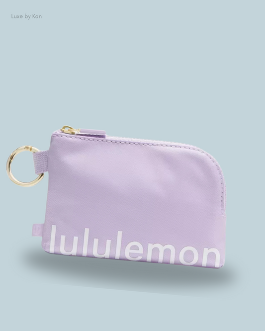 A small lavender zippered pouch with a gold keyring, featuring the word "lululemon" in white. Set against a soft blue background.