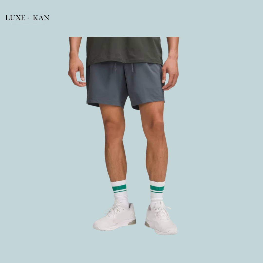 A man stands wearing gray shorts, white sneakers, and green-striped socks, showcasing a casual athletic look against a light blue background.