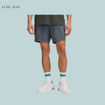 A man stands wearing gray shorts, white sneakers, and green-striped socks, showcasing a casual athletic look against a light blue background.