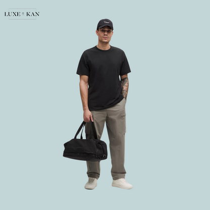 A person stands against a light blue background, wearing a black t-shirt and gray cargo pants, holding a black duffel bag.